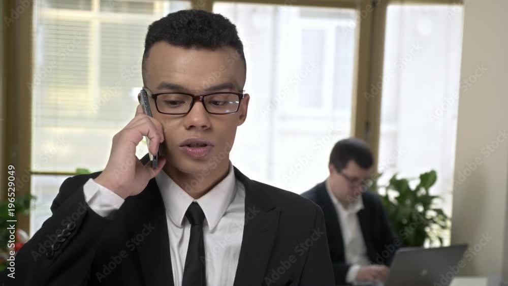 Young successful African businessman talking on a smartphone in the office, Man working at the computer in the background. 60 fps Young successful African businessman talking on a smartphone in the office, Man working at the computer in the background. 60 fps