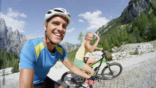 SELFIE: Happy couple on bicycle ride smiles as fit young woman waves to camera.