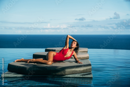 Beautiful view of sexy girl relaxing at swimming pool with ocean at background. Lovely young woman in red bikini posing. Concept of swimsuit, swimwear and travel