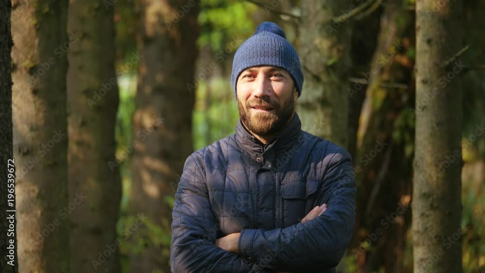 Happy smiling young bearded guy crossing his arms looking at camera in city park. clothes jacket and knitted hat. Happy smiling young bearded guy crossing his arms looking at camera in city park. clothes jacket and knitted hat.