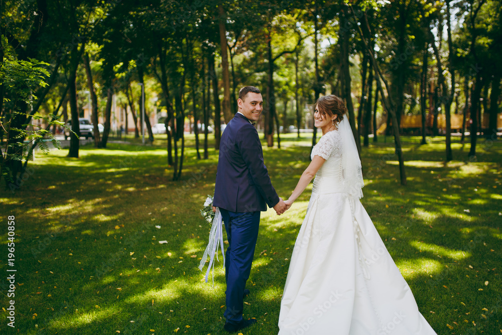 Beautiful wedding photosession. Handsome groom in blue formal suit with bouquet and his elegant bride in white dress and veil with beautiful hairdress on a walk in the big green park on a sunny day