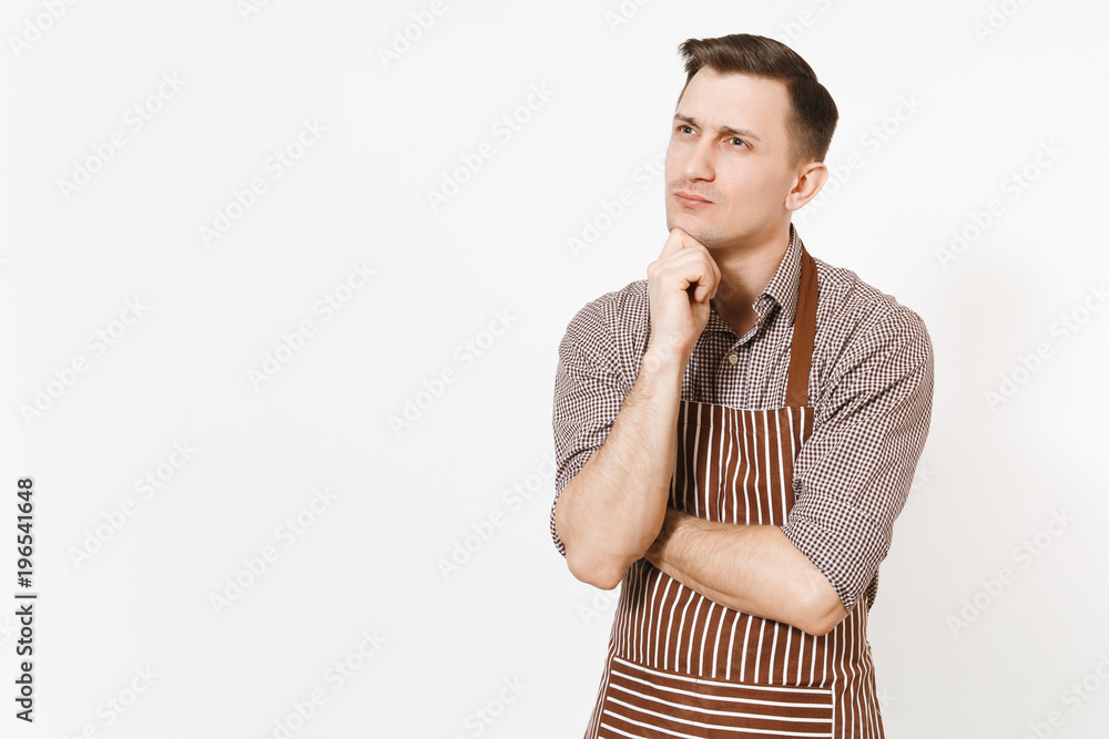 Young pensive man chef or waiter in striped brown apron, shirt holding hand to chin isolated on white background. Male housekeeper or houseworker - looking camera. Domestic worker for advertisement. Young pensive man chef or waiter in striped brown apron, shirt holding hand to chin isolated on white background. Male housekeeper or houseworker - looking camera. Domestic worker for advertisement.
