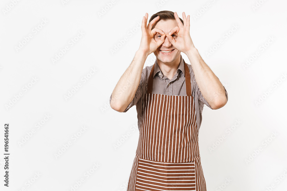 Young smiling man chef or waiter in striped brown apron, shirt holding hands near eyes, imitating glasses or binoculars isolated on white background. Male housekeeper or houseworker. Domestic worker. Young smiling man chef or waiter in striped brown apron, shirt holding hands near eyes, imitating glasses or binoculars isolated on white background. Male housekeeper or houseworker. Domestic worker.