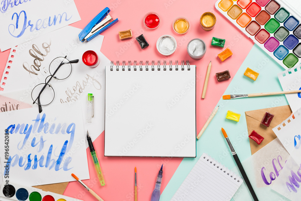 Beautiful flatlay arrangement with watercolors, brushes, glasses, brushpen, paints with notebook with blank copyspace page and other stationary and art supplies. Creativity or education concept Beautiful flatlay arrangement with watercolors, brushes, glasses, brushpen, paints with notebook with blank copyspace page and other stationary and art supplies. Creativity or education concept