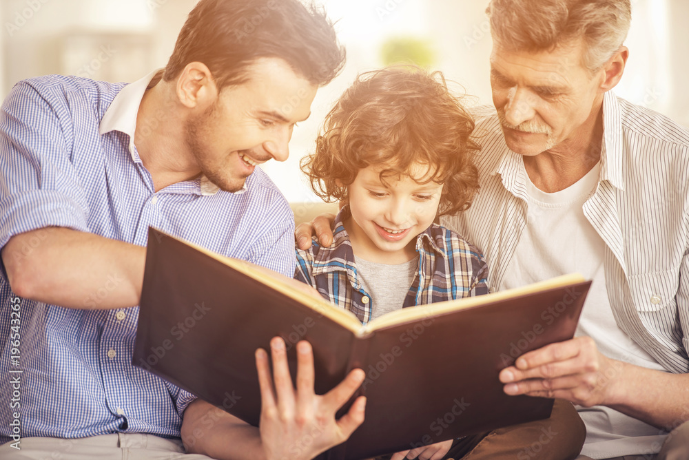 Generation portrait. Grandfather, father and son sitting and reading a book on sofa Generation portrait. Grandfather, father and son sitting and reading a book on sofa