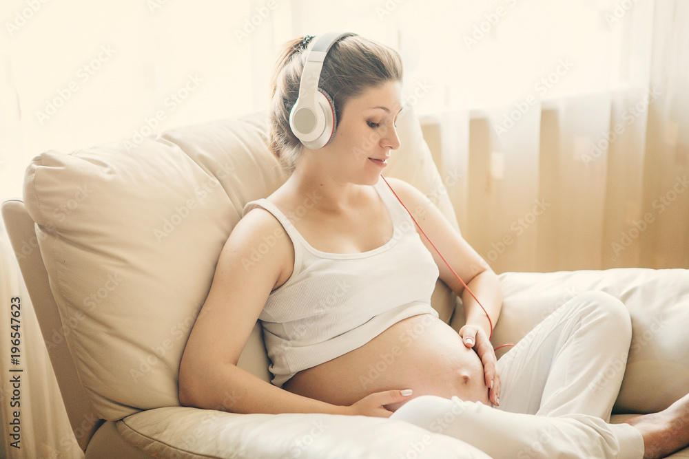 Portrait of pregnant woman sitting on sofa at home and listening music in headphones.