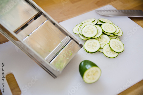 A pile of zucchini slices sits on a cutting board next to a mandolin photo