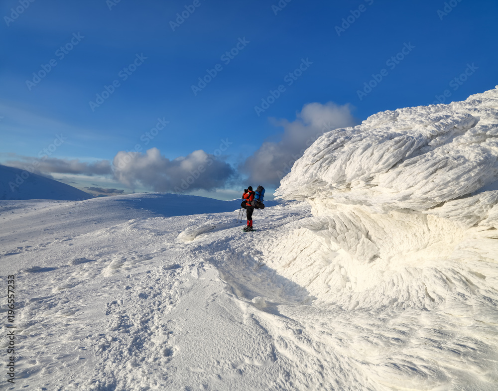 A panorama of snow capped mountains with textured, frozen, mystical fanciful shaped stones, a man in a ski suit with a backpack and a camera.