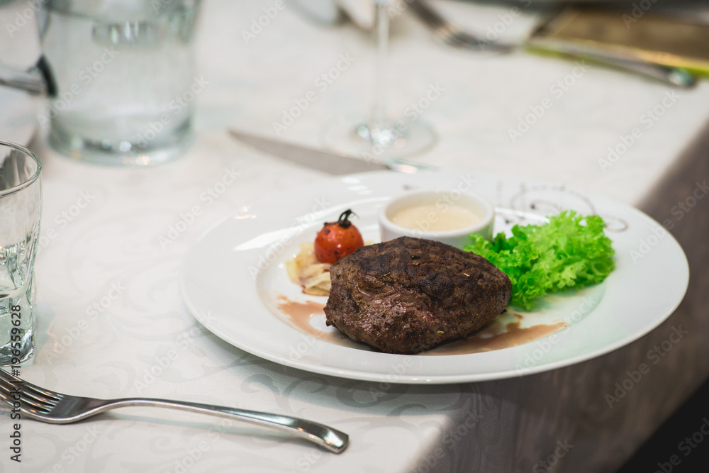 fresh grilled bbq roast beef steak and sauce on a white plate with green leaf of salad. soup sauce small jug glass served on a table in a restaurant