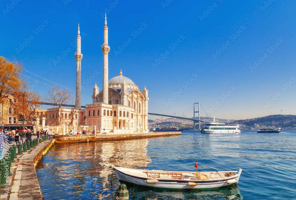 Ortakoy cami - famous and popular landmark in Istanbul, Turkey. Lovely spring scenery with fishing boat at foreground and old historical mosque Ortakoy and Istanbul Bosporus bridge at background. - obrazy, fototapety, plakaty 