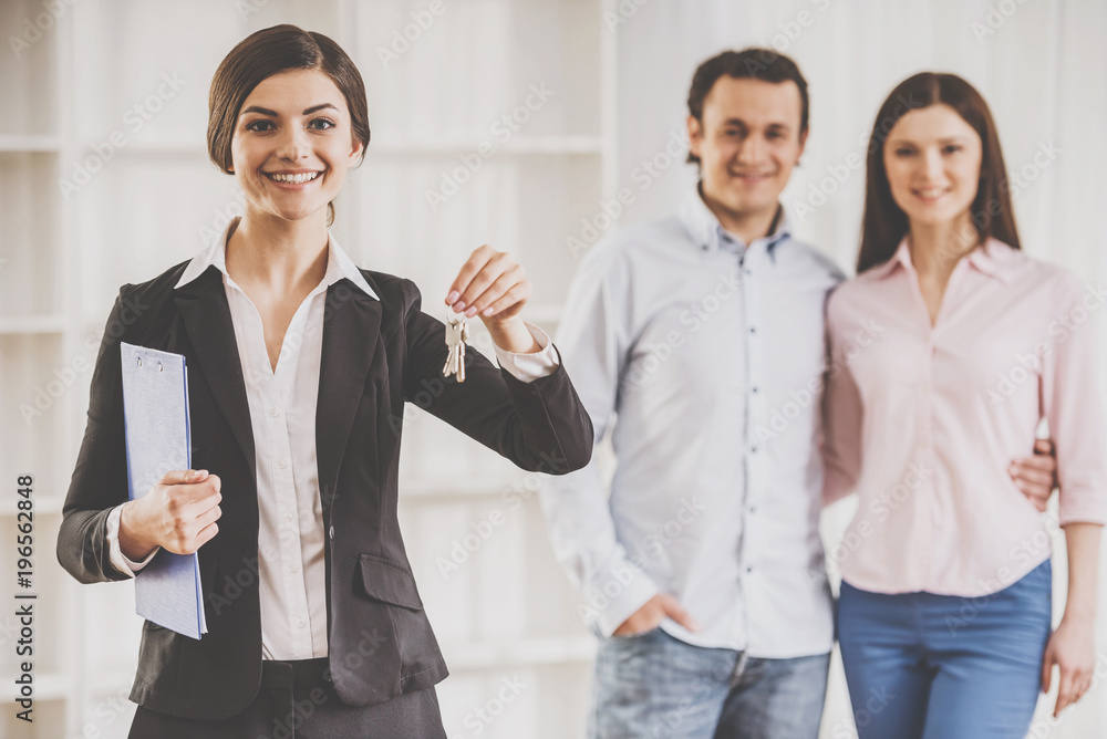 Realtor woman is holding keys and young couple on background Realtor woman is holding keys and young couple on background