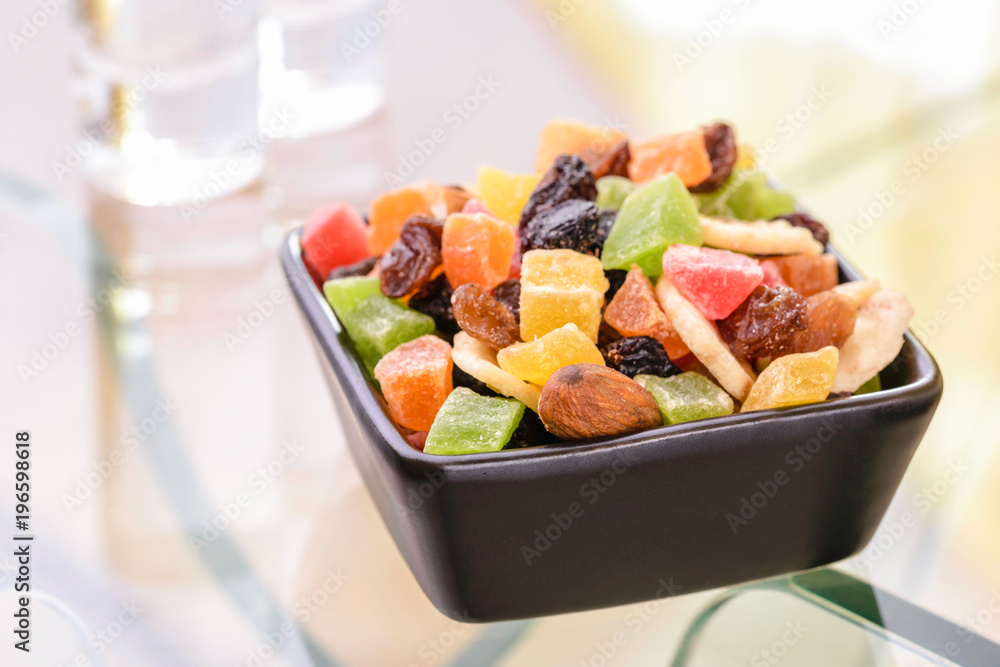 Many colorful dry fruits in a little black square bowl put on a glass table, with the reflection of two vodka glasses Many colorful dry fruits in a little black square bowl put on a glass table, with the reflection of two vodka glasses