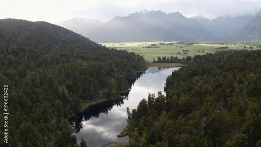 Wide drone shot of a clear reflective lake surrounded by trees and snow-capped mountains. Wide drone shot of a clear reflective lake surrounded by trees and snow-capped mountains.