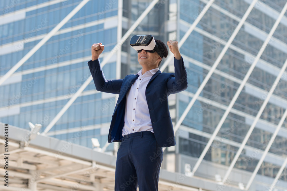 Young Asian businessman in a suit wear and virtual reality glasses. He are glad that succeed with city town background. Concept Technology and Simulation Business.. Young Asian businessman in a suit wear and virtual reality glasses. He are glad that succeed with city town background. Concept Technology and Simulation Business..
