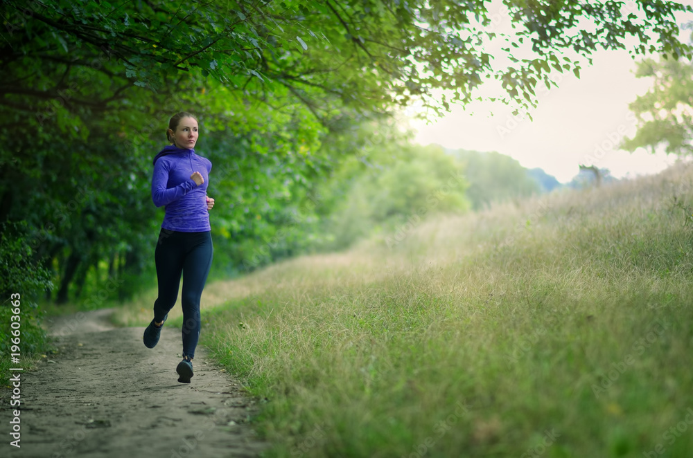 young slim runner in a black sports leggins and and blue jacket fast runs along the path on the beautiful green forest. young slim runner in a black sports leggins and and blue jacket fast runs along the path on the beautiful green forest.