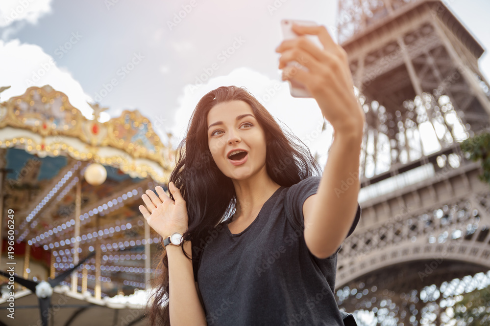 Happy travel woman taking funny selfie with her mobile phone near the Eiffel tower and carousel, Paris. Portrait of travel tourist girl on vacation walking happy outdoors. Attractive young romantic