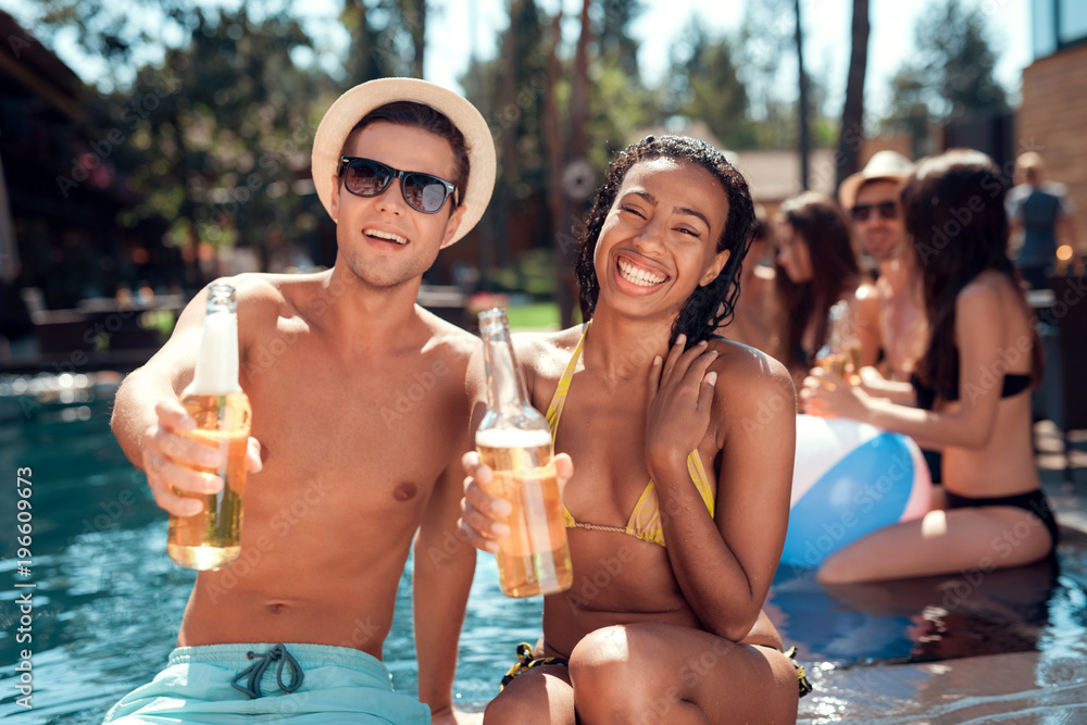 Young man is sunglasses and cheerful woman cheering with beer bottles in swimming pool.