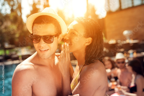 Fotografie Young black girl in swimsuit gossiping with young handsome guy in straw hat near pool