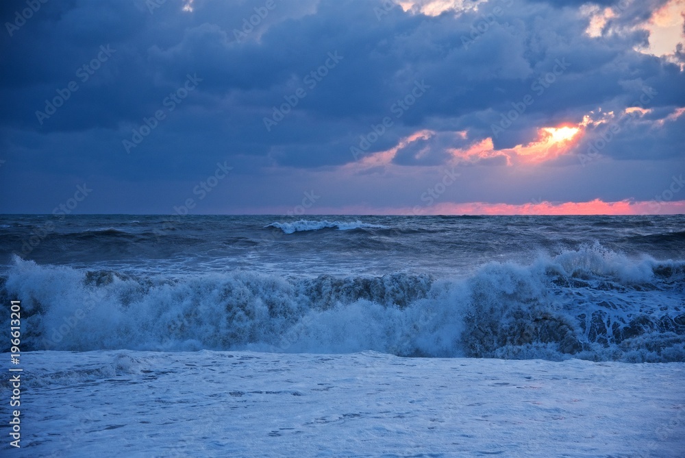 Storm on the sea. Large waves roll on the shore. The sun at sunset makes its way through the clouds.