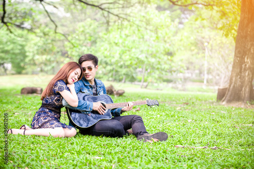Asian couple in love playing acoustic song guitar sitting on grass in the park