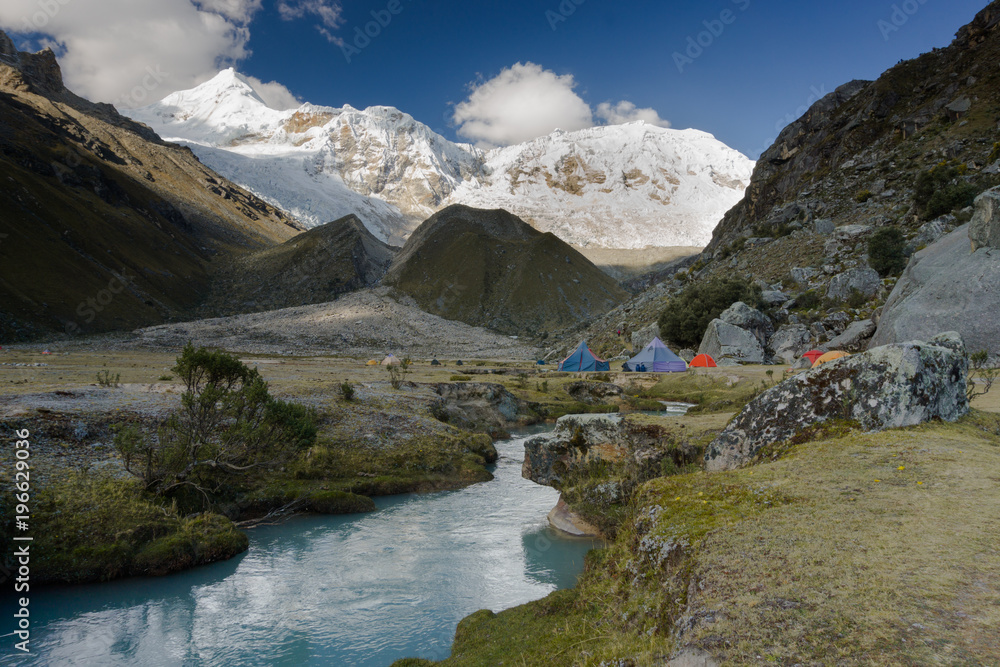 base camp in the Andes in Peru with snowy high peaks behind and large boulders and red rocks and a little mountain stream in the foreground