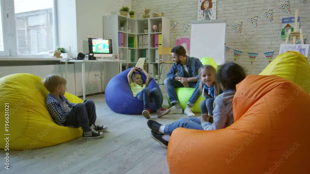 Preschool boys and girls sitting on colorful bean bags chairs around male teacher and speaking with him while taking a lesson in kindergarten classroom Preschool boys and girls sitting on colorful bean bags chairs around male teacher and speaking with him while taking a lesson in kindergarten classroom