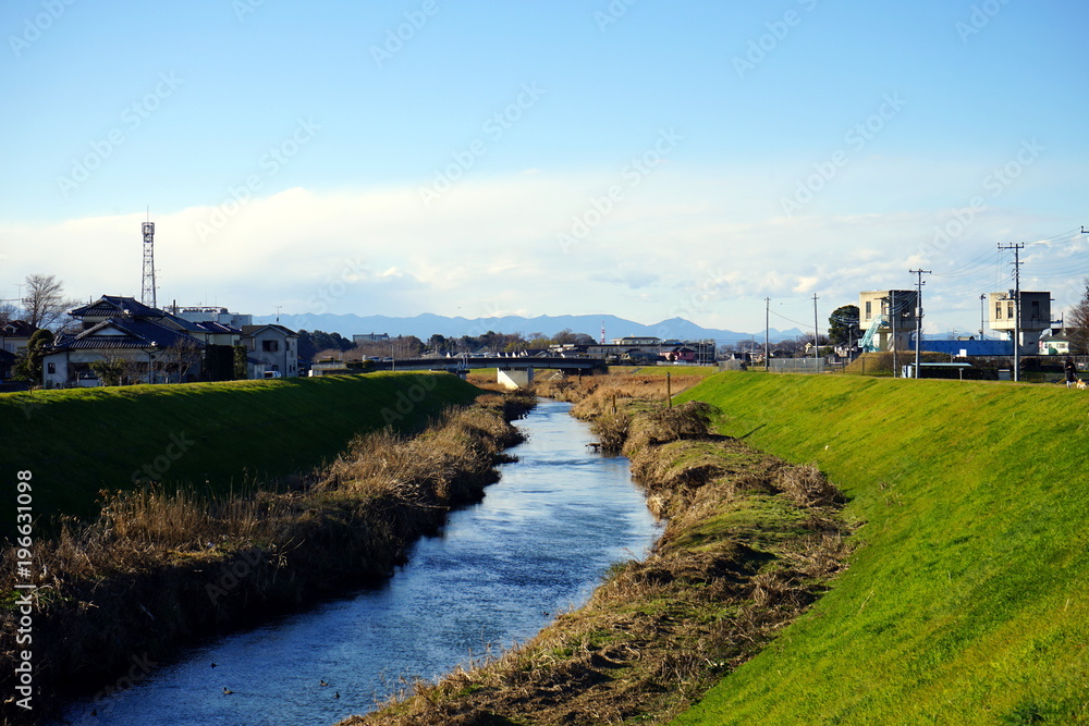 A landscape of river with green grass, small house, and background mountain in suburban under the blue sky A landscape of river with green grass, small house, and background mountain in suburban under the blue sky