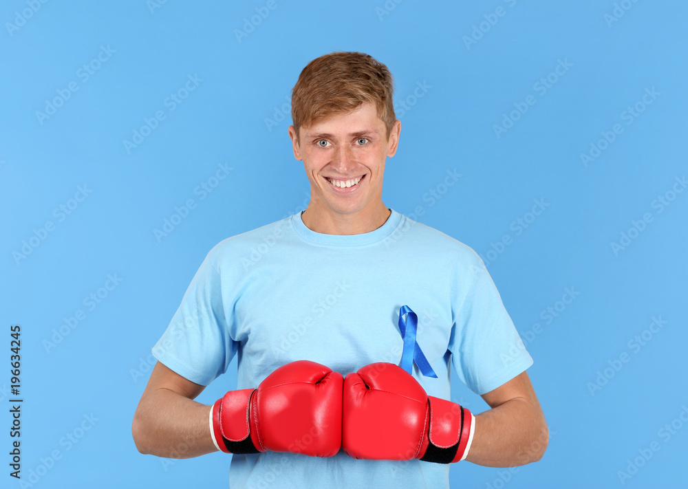 Young man in t-shirt with blue ribbon wearing boxing gloves on color background. Prostate cancer awareness concept Young man in t-shirt with blue ribbon wearing boxing gloves on color background. Prostate cancer awareness concept