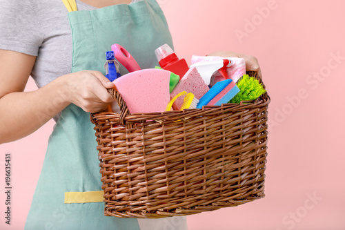 Woman holding basket with cleaning supplies on color background