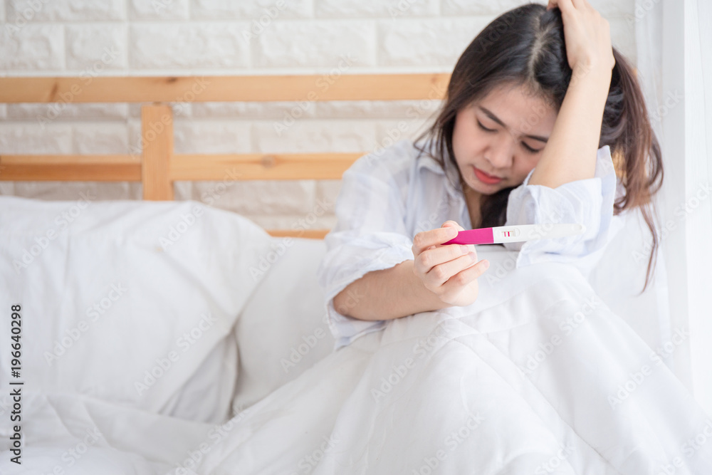 Selective focus Pregnancy test positive result on hand of woman sitting on bed in her bedroom. Pregnancy is not ready concept. Selective focus Pregnancy test positive result on hand of woman sitting on bed in her bedroom. Pregnancy is not ready concept.