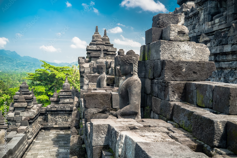 Amazing view of ancient Borobudur Buddhist temple with meditating Buddha statue carved from dark stone against blue sky on background. Great religious architecture. Magelang, Central Java, Indonesia