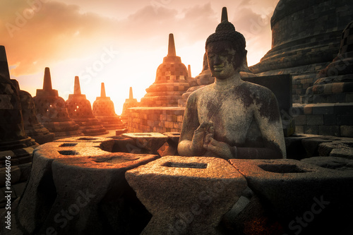 Amazing sunrise view of meditating Buddha statue and stone stupas against shining sun on background. Ancient Borobudur Buddhist temple. Great religious architecture. Magelang, Central Java, Indonesia © PerfectLazybones