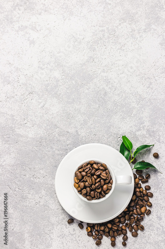 Coffee beans in white cup and green leaves on concrete background. Top view, space for text.
