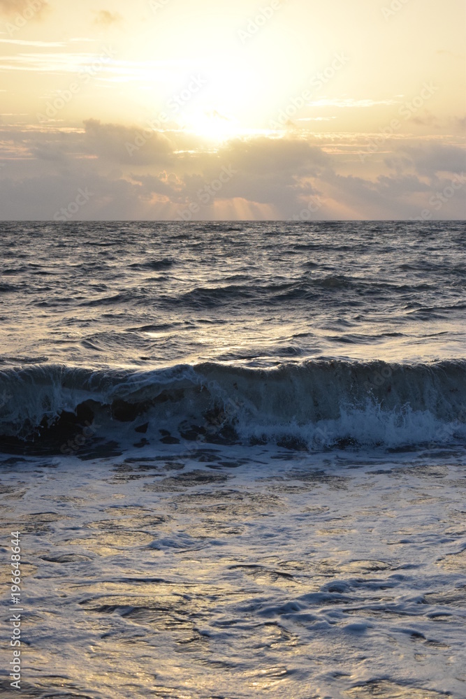 Ocean at sunrise with wave crashing on beach, clouds orange sky.