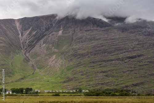 Annat, Scotland - June 10, 2012: Massive brown-green mountain wall with top in clouds. Fine line of white houses at its base partly hidden by green trees. Meadow up front.