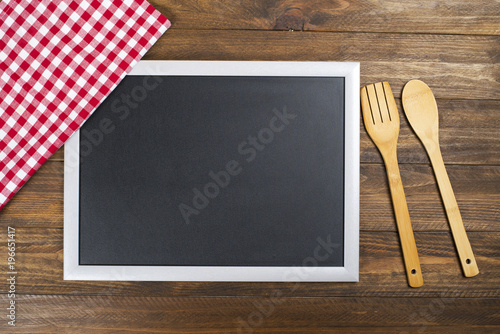 Blackboard on brown wooden table next to red and white cloth napkin and wooden spoon and fork.
