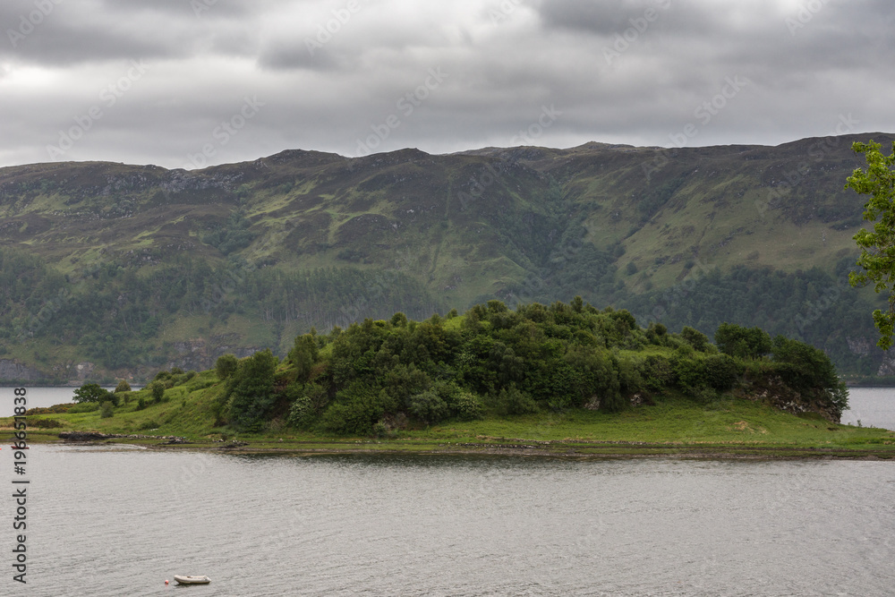 Lochcarron, Scotland - June 10, 2012: Green vegetation on island in Loch Carron. Gray water and rain-heavy cloudscape. Forested Mountain wall on opposite shore.