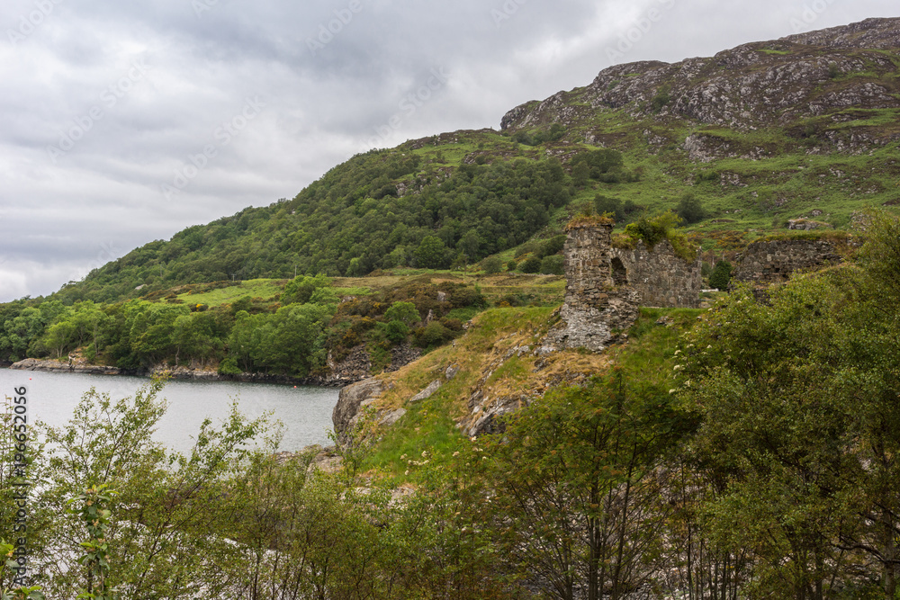 Stromeferry, Scotland - June 10, 2012: Castle Strome ruins on green shore cliff over Loch Carron. Cloudscape and forested mountain in back. Top of trees in front.