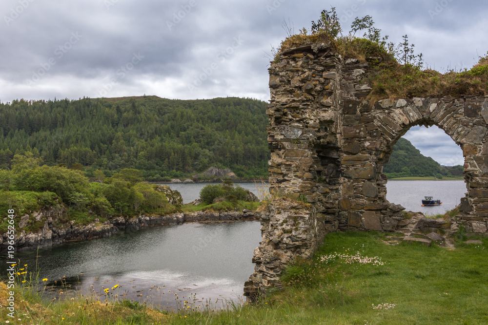 Stromeferry, Scotland - June 10, 2012: Window in rock wall of Castle Strome ruins on green hill. Cloudscape and Loch Carron with motor boat seen in back. Mountains on opposite shore and horizon.
