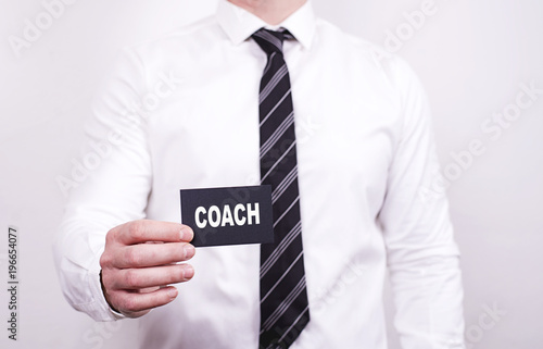 Man with a black tie and white shirt holds in one hand a card on which the word Coach is written. Business