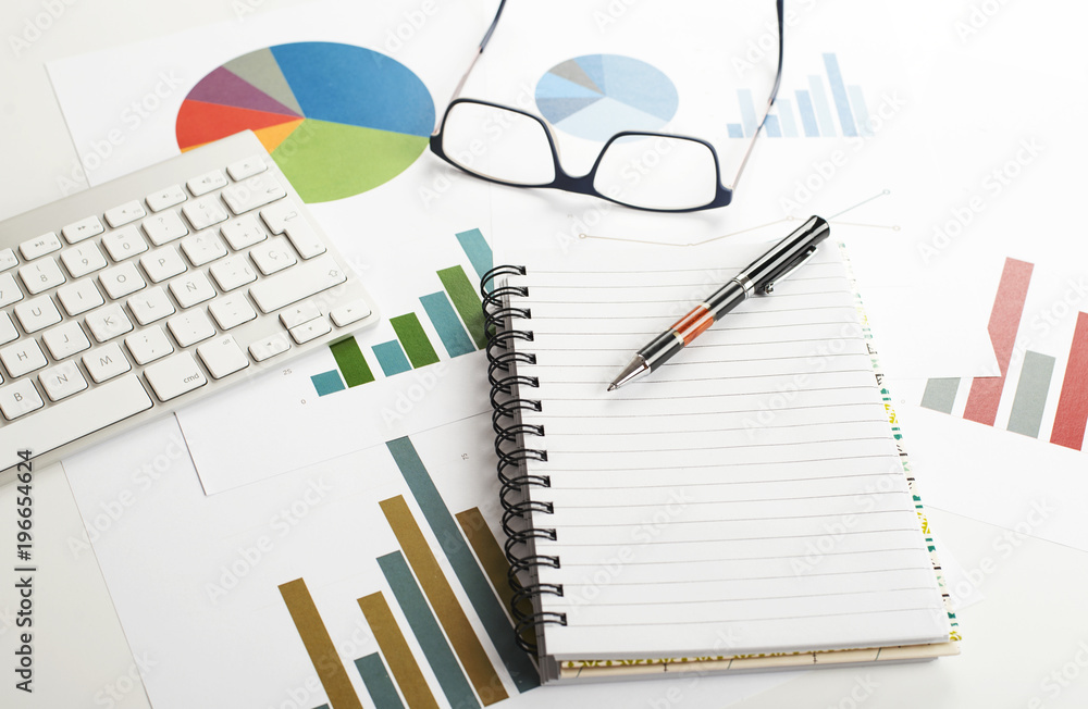 Background top view of workplace with economic graphics of various colors next to keyboard, pen and glasses with an open notebook in the middle of the image. Copy space. Mockup. Background top view of workplace with economic graphics of various colors next to keyboard, pen and glasses with an open notebook in the middle of the image. Copy space. Mockup.