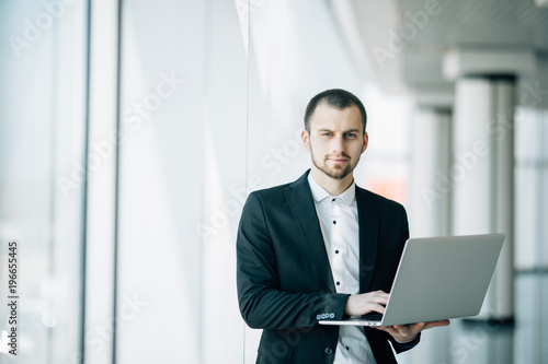 Businessman stands in a bright airy office holding an open laptop in his hands
