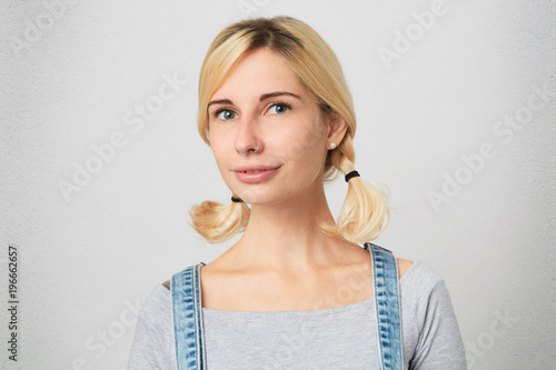 Indoor shot of young blonde girl with perced nose, wears overalls, simply hairdo smiling and show her teeth to the world. Isolated over white concrete wall photo