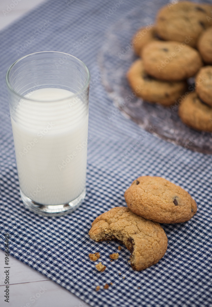 A glass of milk and a cookie with a bite taken out of it on a blue gingham napkin with a plate of cookies in the background A glass of milk and a cookie with a bite taken out of it on a blue gingham napkin with a plate of cookies in the background