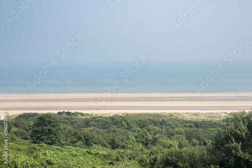 A broad, sandy beach on a bright summers day. The sky is deep blue and hazy from the sunshine. The coastline is Normandy, in Northern France