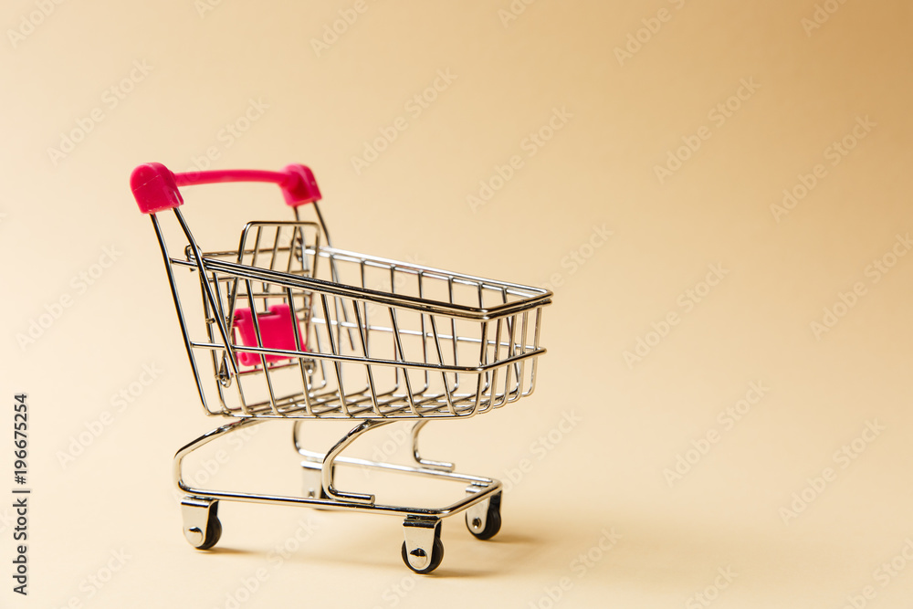 Close up of supermarket grocery push cart for shopping with black wheels and pink plastic elements on handle isolated on beige background. Concept of shopping. Copy space for advertisement Close up of supermarket grocery push cart for shopping with black wheels and pink plastic elements on handle isolated on beige background. Concept of shopping. Copy space for advertisement
