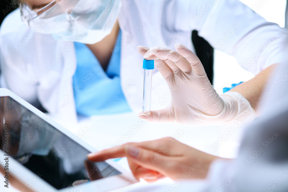 Closeup of scientific research team with clear solution in laboratory. Blonde female chemist holds test tube of glass while her colleague checks results with tablet pc. Blood test, medicine or Closeup of scientific research team with clear solution in laboratory. Blonde female chemist holds test tube of glass while her colleague checks results with tablet pc. Blood test, medicine or