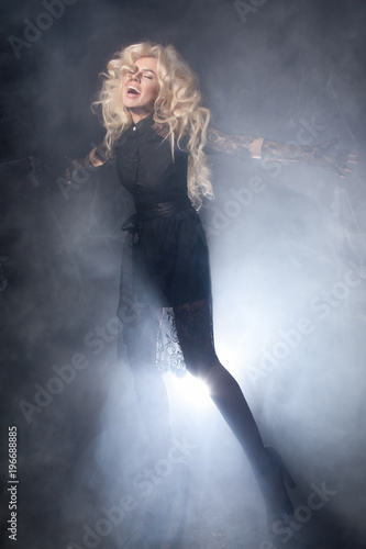 luxurious blonde with a fluffy white long hair and long legs posing in a short black lace dress on a dark stage background in the smoke with the light © goldeneden