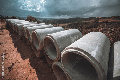 Wide-angle shot with the two rows of concrete or cement constructional pipes laying one on one, which used for the creation of sewerage systems and drain shafts; hills behind and sun rays photo