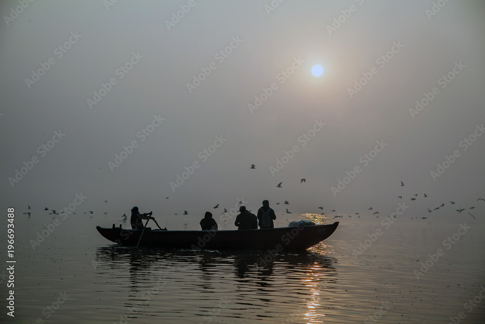 Boat with tourists inside on Ganga river in the morning sunrise in the fog with birds flying on the background, man wearing a backpack, boatman is rowing
